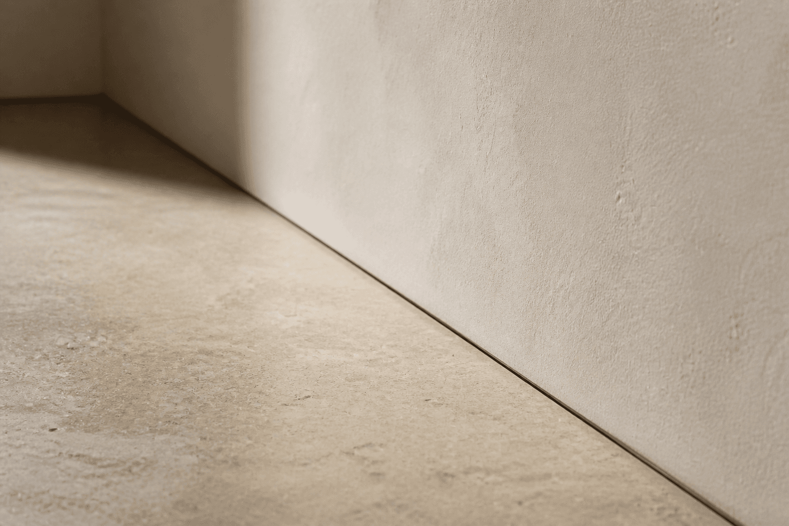 Close study of a honed stone floor meeting a plaster skirting, with a clean shadow gap and no decorative trim